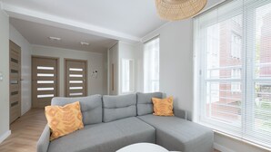Un salon moderne avec un canapé d’angle gris, deux coussins décoratifs jaunes à motifs, une table basse blanche et de grandes fenêtres équipées de stores.