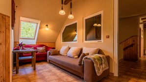 A cozy living room with a brown sofa, a small wooden table, and a red couch. There are two windows with white frames and a wooden staircase in the background. - Ostsee Ferienhaus Welzin - Schulgartenhaus (Damshagen)