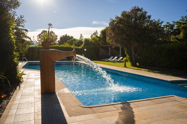 Une piscine agrémentée d’un élément décoratif aquatique, entourée d’une terrasse carrelée et de verdure.