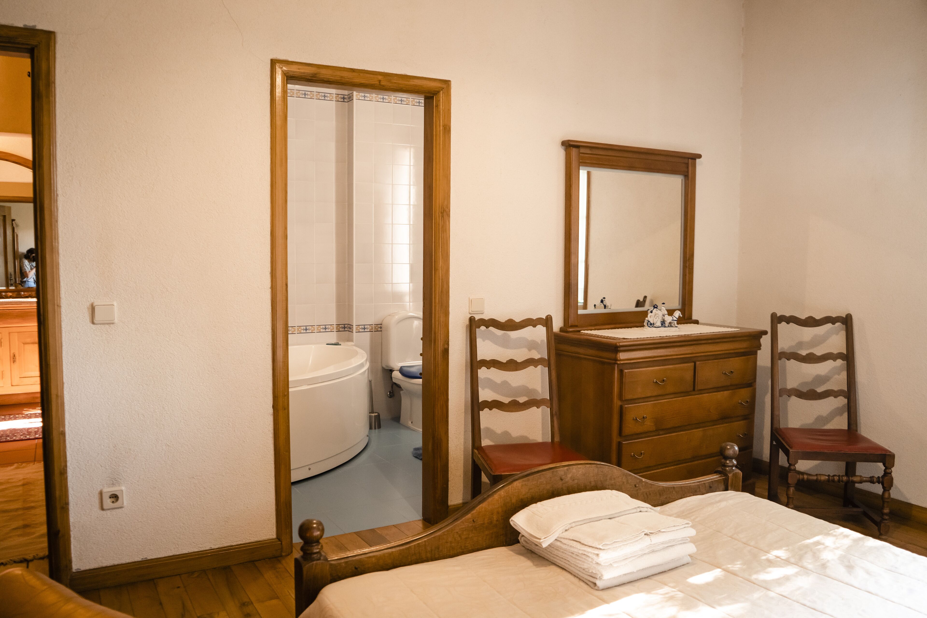 A bedroom with a bed, wooden dresser, two chairs, a mirror, and a bathroom visible through an open door.