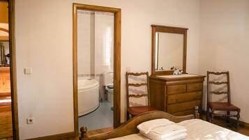 A bedroom with a bed, wooden dresser, two chairs, a mirror, and a bathroom visible through an open door.
