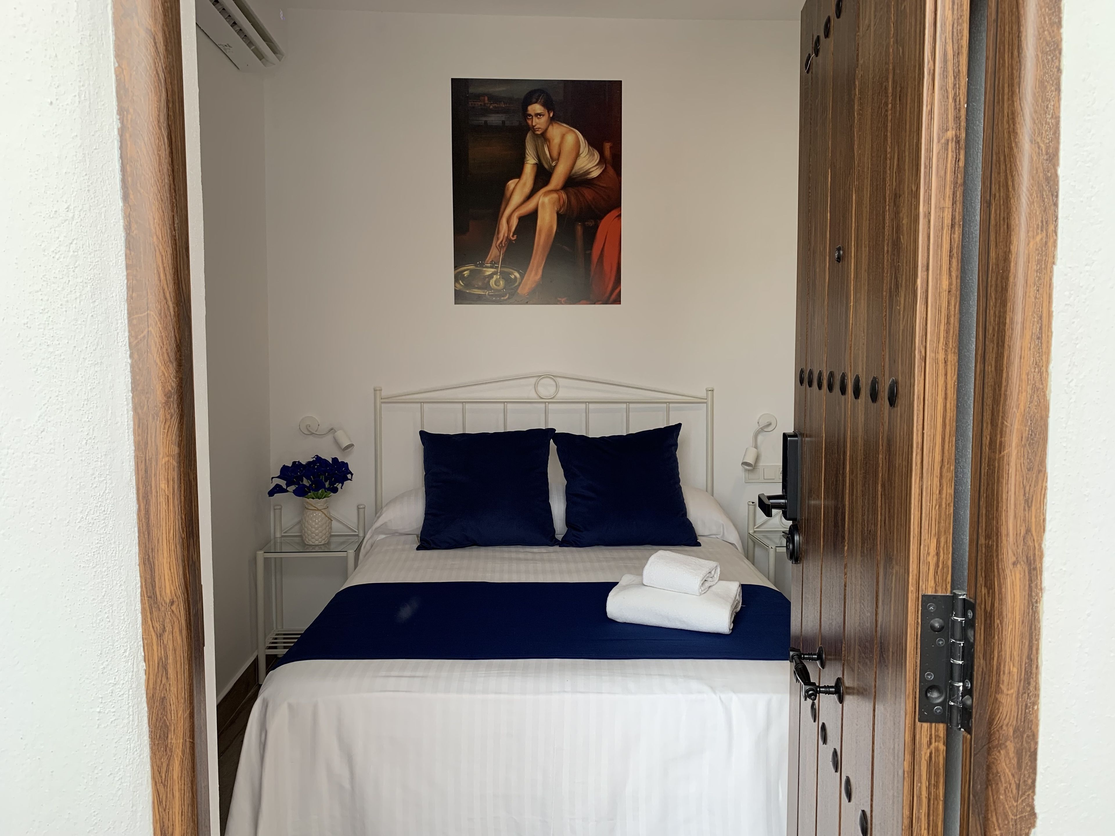 Een net opgemaakt bed met een blauwe deken en kussens, een witte nachtkastje met een bloevas, en een schilderij aan de muur.