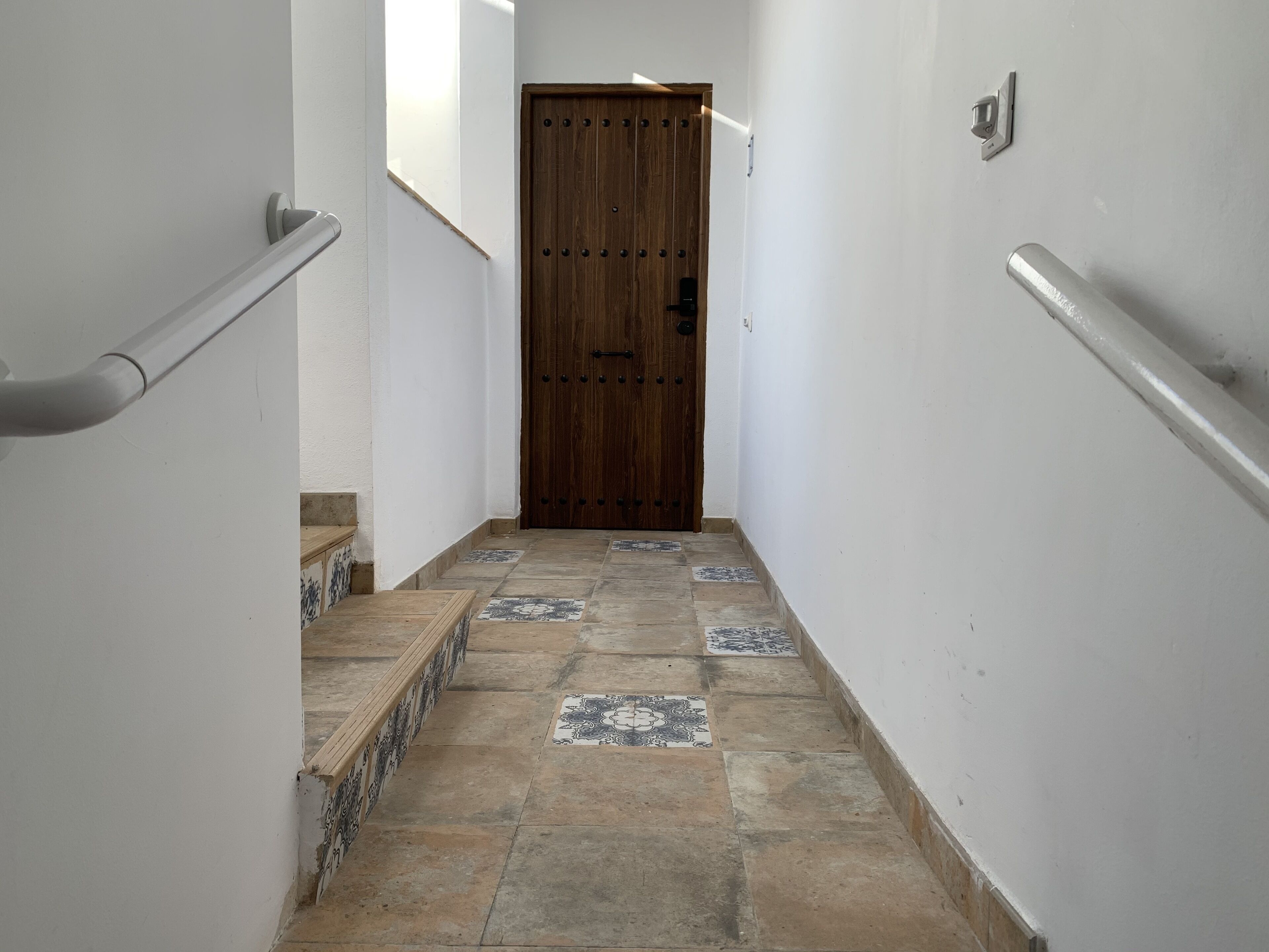 Um corredor estreito com uma porta de madeira no final, piso de azulejos e paredes brancas.