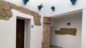Ein schmaler Gang mit einer Holztür, einer Ziegelstütze und einer weißen Wand mit einem Ziegelbereich. An der Wand befinden sich blaue Blumentöpfe und eine kleine Wandleuchte.