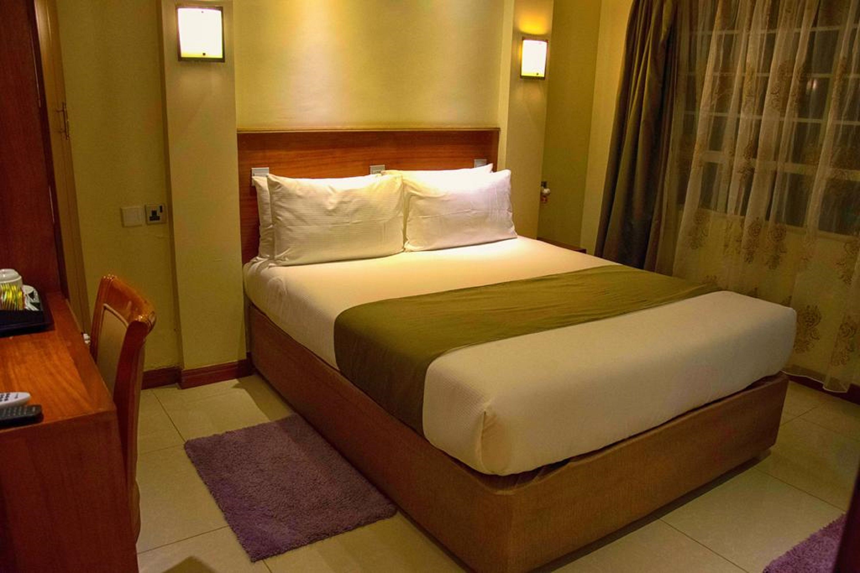 Une chambre d’hôtel avec un grand lit, une tête de lit en bois, deux lampes de chevet, un bureau avec une chaise et un tapis violet.