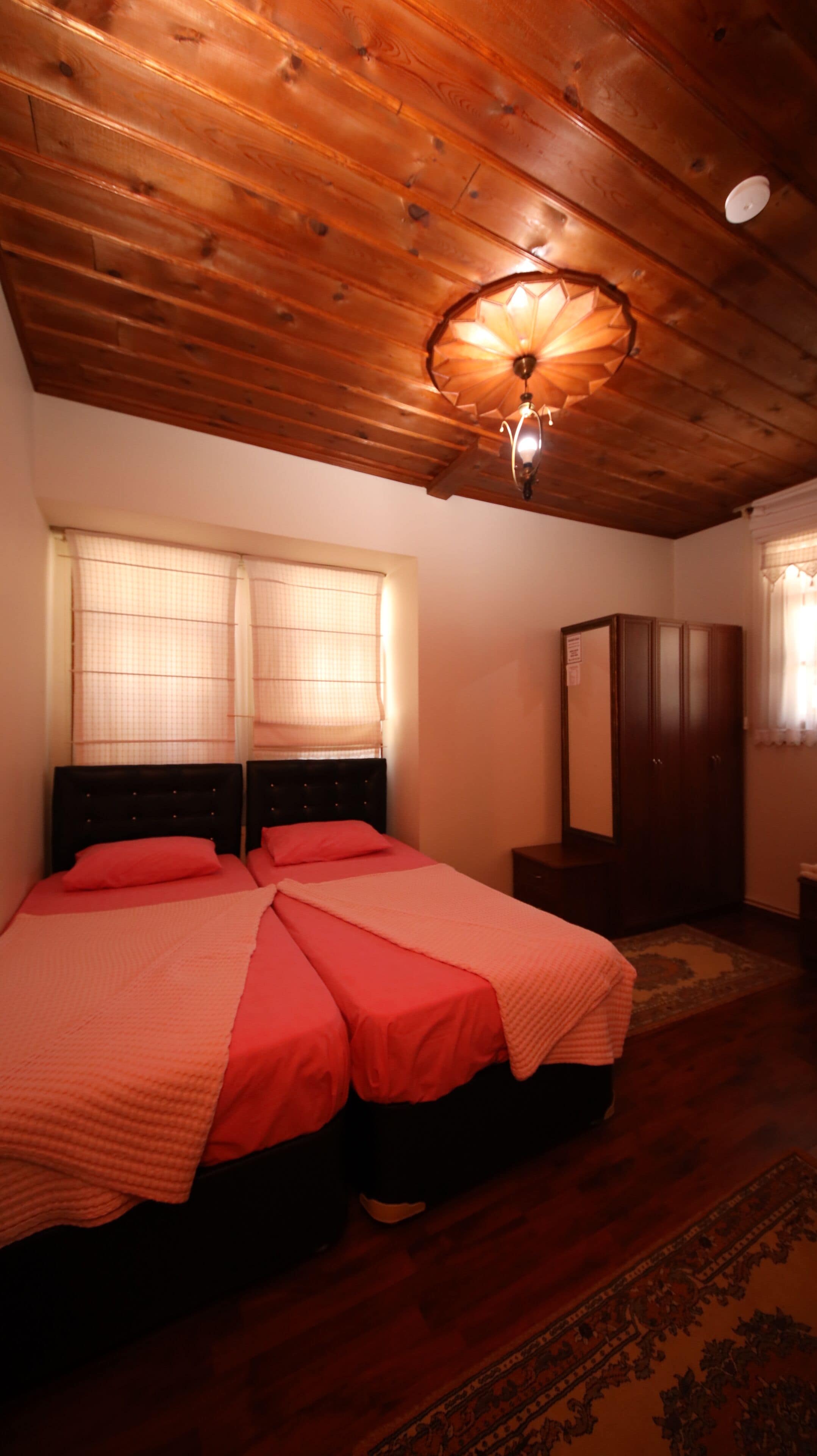 A bedroom with a wooden ceiling, two beds with red bedding, a nightstand, a wardrobe, and a patterned rug.