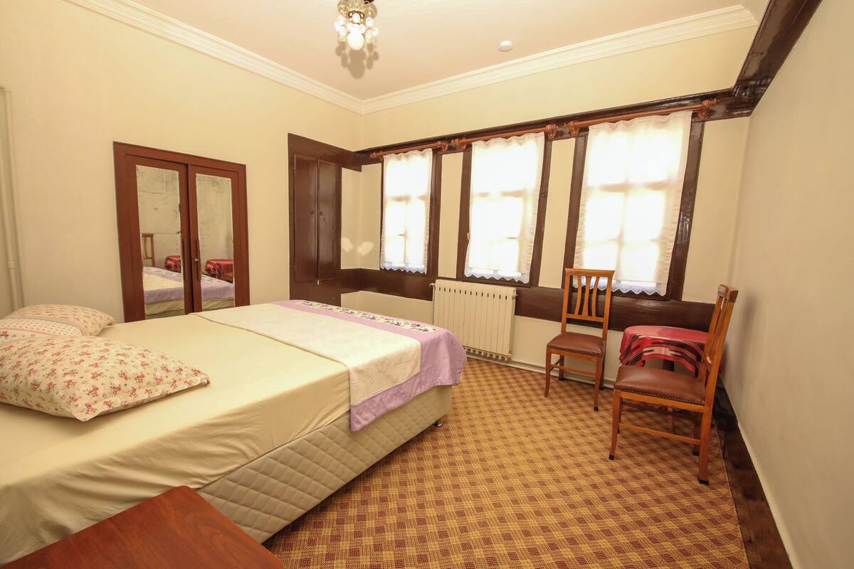 Une chambre à coucher avec un lit, une chaise, une table, un miroir et une fenêtre avec des rideaux.