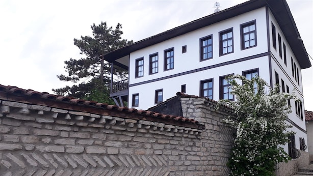 Un edificio de dos plantas con un muro de piedra al frente y una fachada blanca con detalles oscuros.