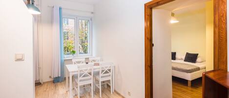 Una habitación pequeña y luminosa con piso de madera, una mesa blanca con sillas, una ventana con cortinas y una puerta que da a otra habitación con una cama.