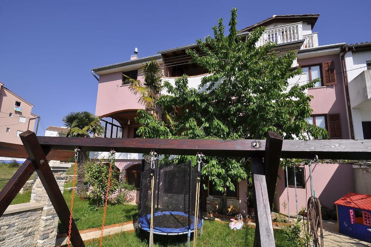 A residential area with a trampoline in the yard, surrounded by greenery and a pink building in the background.