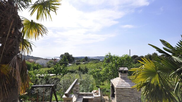 A patio area with a stone barbecue and a swing set, surrounded by lush greenery and a view of a distant landscape. - Apartments Margareta (Pula)