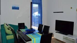 En kompakt opholdsstue med spiseplads, sofa, fjernsyn og et vindue med blå gardiner.
