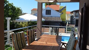 En balkon med et træbord og stribede stole, med udsigt over en swimmingpool og tilstødende bygninger.