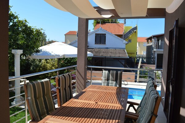 A balcony with a wooden table and striped chairs, overlooking a swimming pool and neighboring buildings.