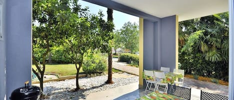 An outdoor patio area with a barbecue grill, a table with chairs, and a view of a garden with trees and a pathway.