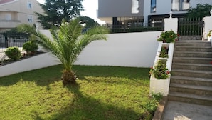 A well-maintained garden with a palm tree, a small flower bed with red flowers, and a concrete staircase leading up to a white wall. - Apartments Adria (Zadar)