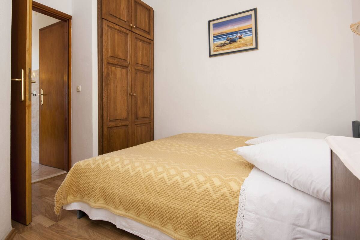 Quarto com uma cama, um guarda-roupa de madeira, um quadro na parede e uma porta que dá para outro cômodo.