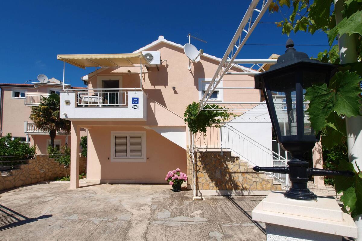 Edifício residencial com varanda, antena parabólica e pequeno jardim com flores cor-de-rosa.