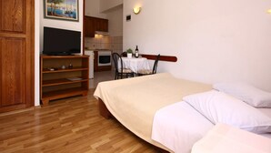 A bedroom with a bed, a TV on a wooden stand, a dining table with chairs, and a kitchen in the background. - Apartments Skarpa (Rogoznica)