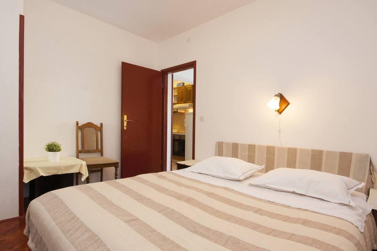 Um quarto com cama, mesa de cabeceira, cadeira, uma pequena mesa com uma planta e porta que dá para uma cozinha.