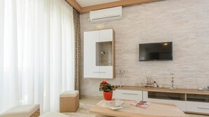 A modern living room with a large flat-screen TV mounted on the wall, a wooden coffee table, a white sofa, and a small side table with a red vase. - Apartments Villa Aurora 3 (Okrug)