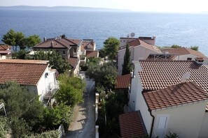 A coastal residential area with terracotta-tiled roofs, a narrow street lined with trees, and a view of the sea in the background.