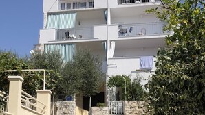 A multi-story residential building with balconies, a stone wall entrance, and a dirt path leading to the building.