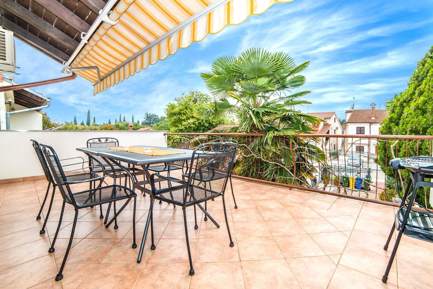 A patio with a table and chairs, a covered area, and a view of surrounding buildings and trees.