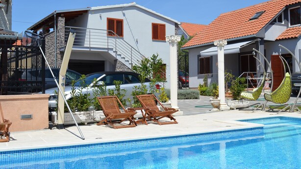 A poolside area with a clear blue pool, wooden lounge chairs, and a two-story building with a red-tiled roof. - Apartments Branko / Joanna / Two Bedrooms (Tribunj)
