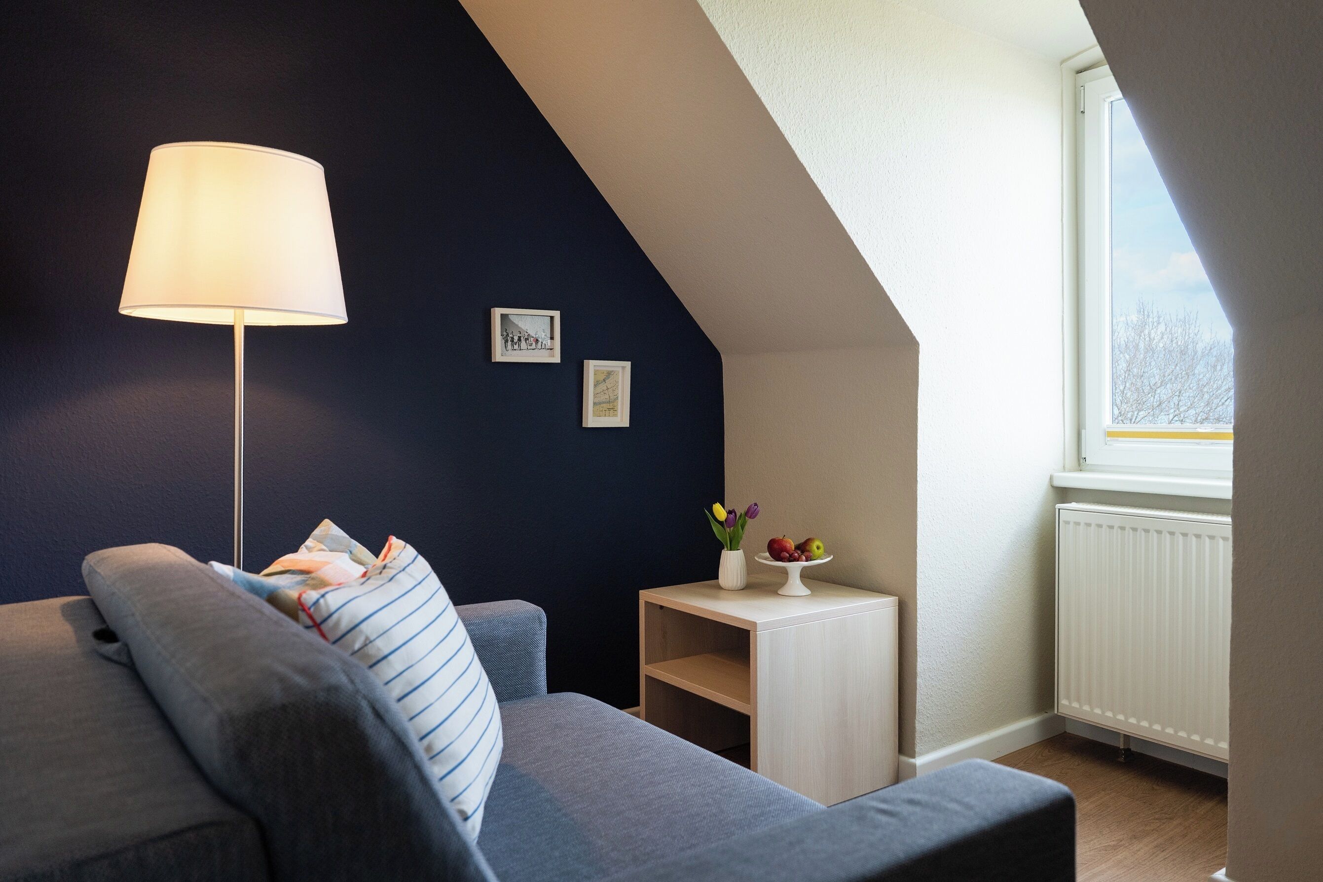 A cozy room with a grey sofa, a floor lamp, a small table with a vase and fruit, and a window with a view.