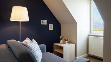 A cozy room with a grey sofa, a floor lamp, a small table with a vase and fruit, and a window with a view.