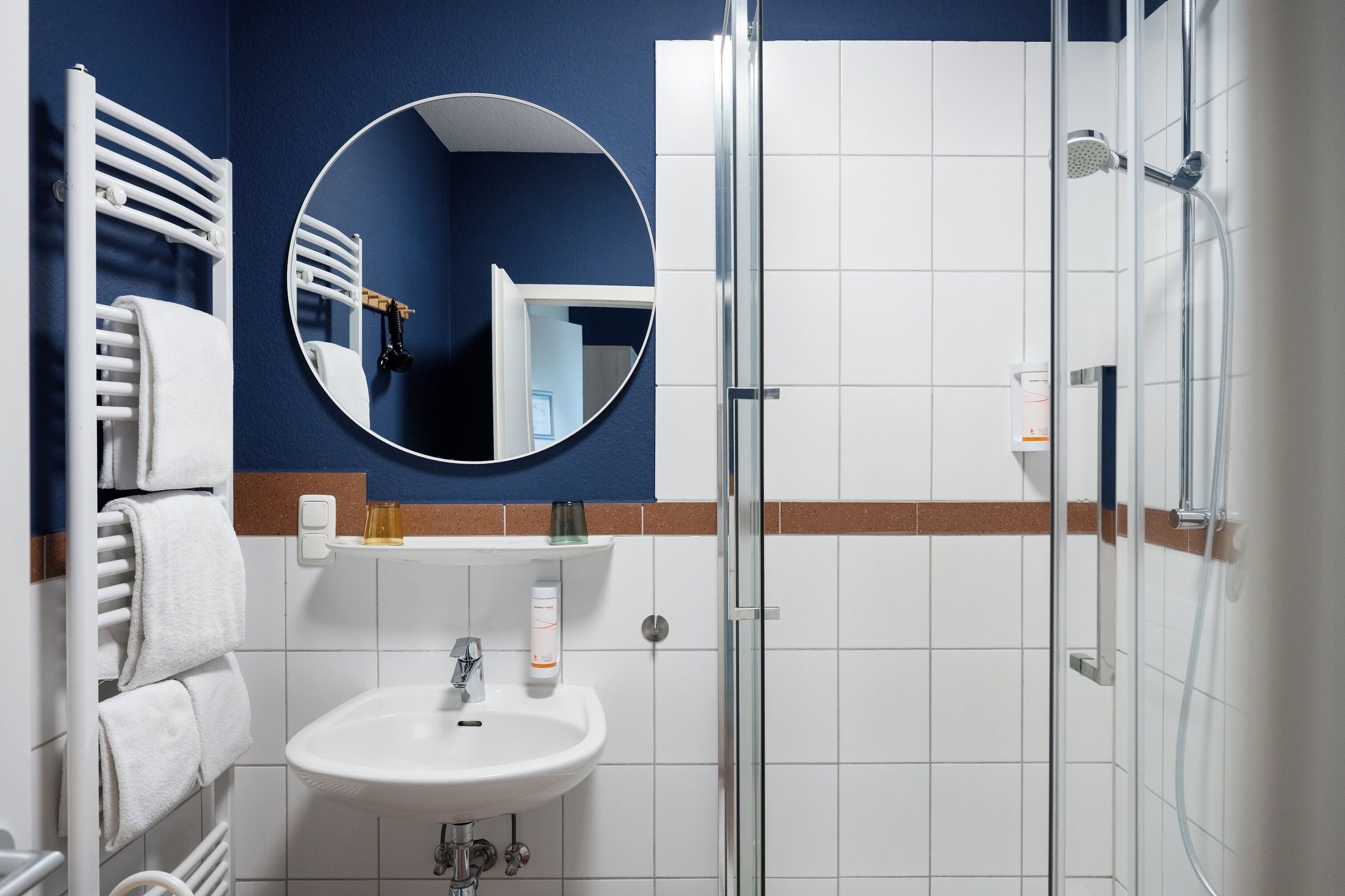 A bathroom with a white sink, a round mirror, a towel rack, and a shower enclosure.