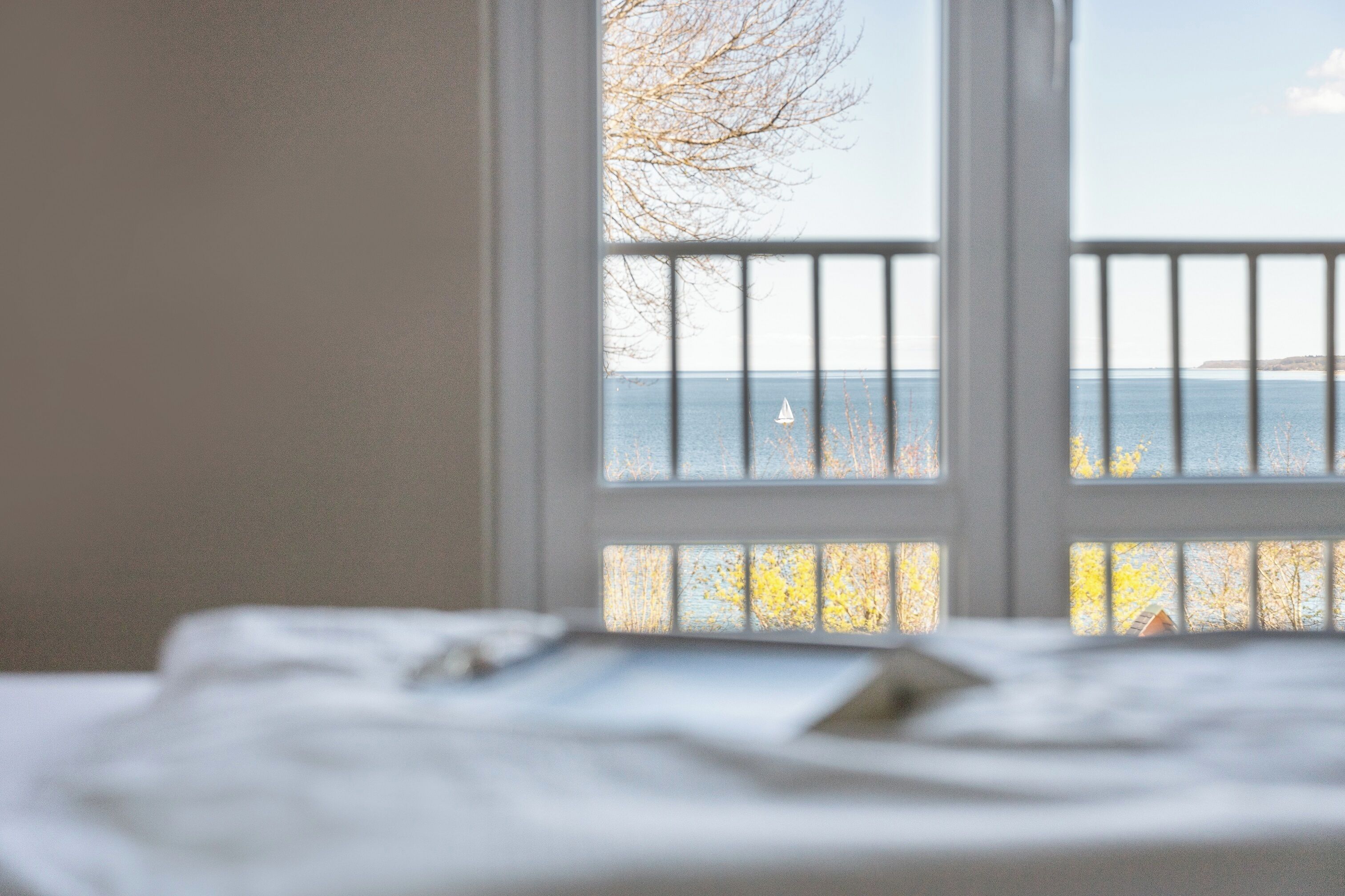 A room with a bed, a window with a view of the sea, and a balcony railing.