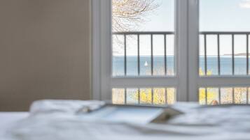 A room with a bed, a window with a view of the sea, and a balcony railing.