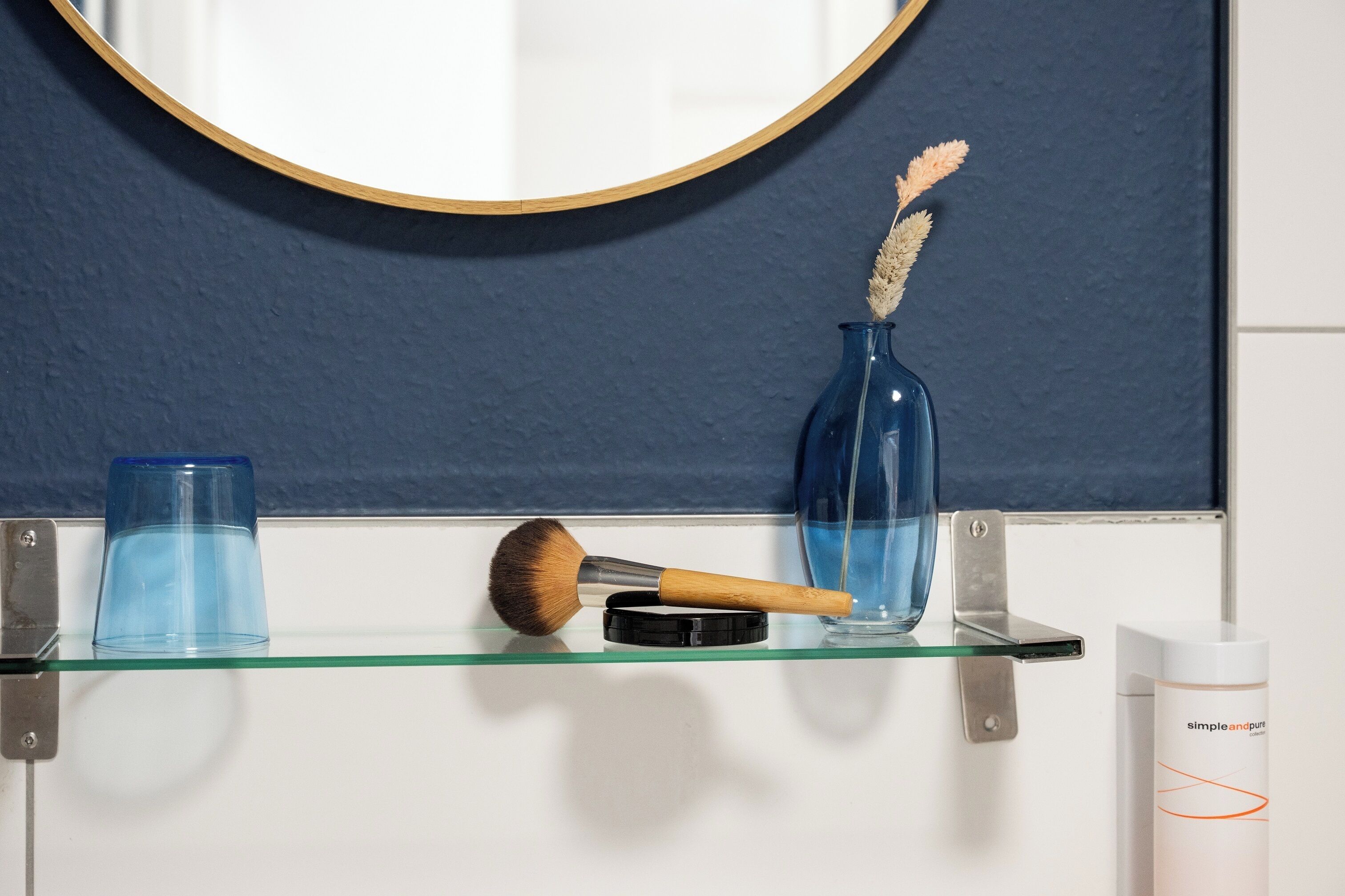 A bathroom with a round mirror, a blue vase with dried plants, a makeup brush, and a glass container on a glass shelf.