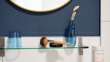 Une salle de bain avec un miroir rond, un vase bleu contenant des plantes séchées, un pinceau à maquillage et un récipient en verre posé sur une étagère en verre.