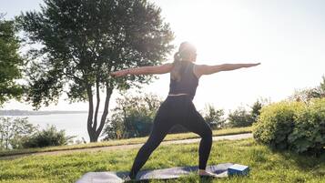 Una persona realizando una postura de yoga sobre una esterilla en un área cubierta de césped, con árboles y un cuerpo de agua al fondo.