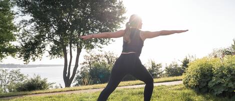 A person performing a yoga pose on a mat in a grassy area with trees and a body of water in the background.
