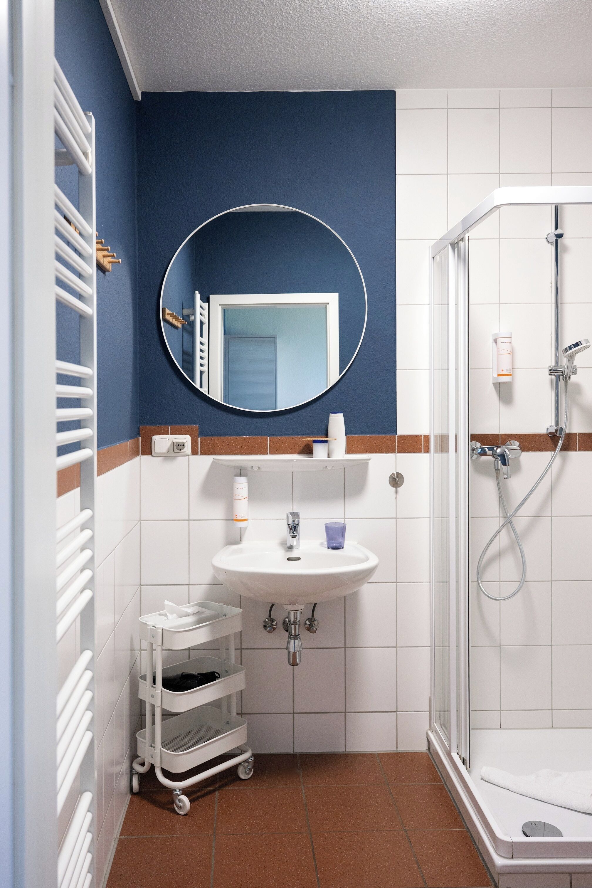 A bathroom with a white sink, a mirror, a shower, and a white storage cart.