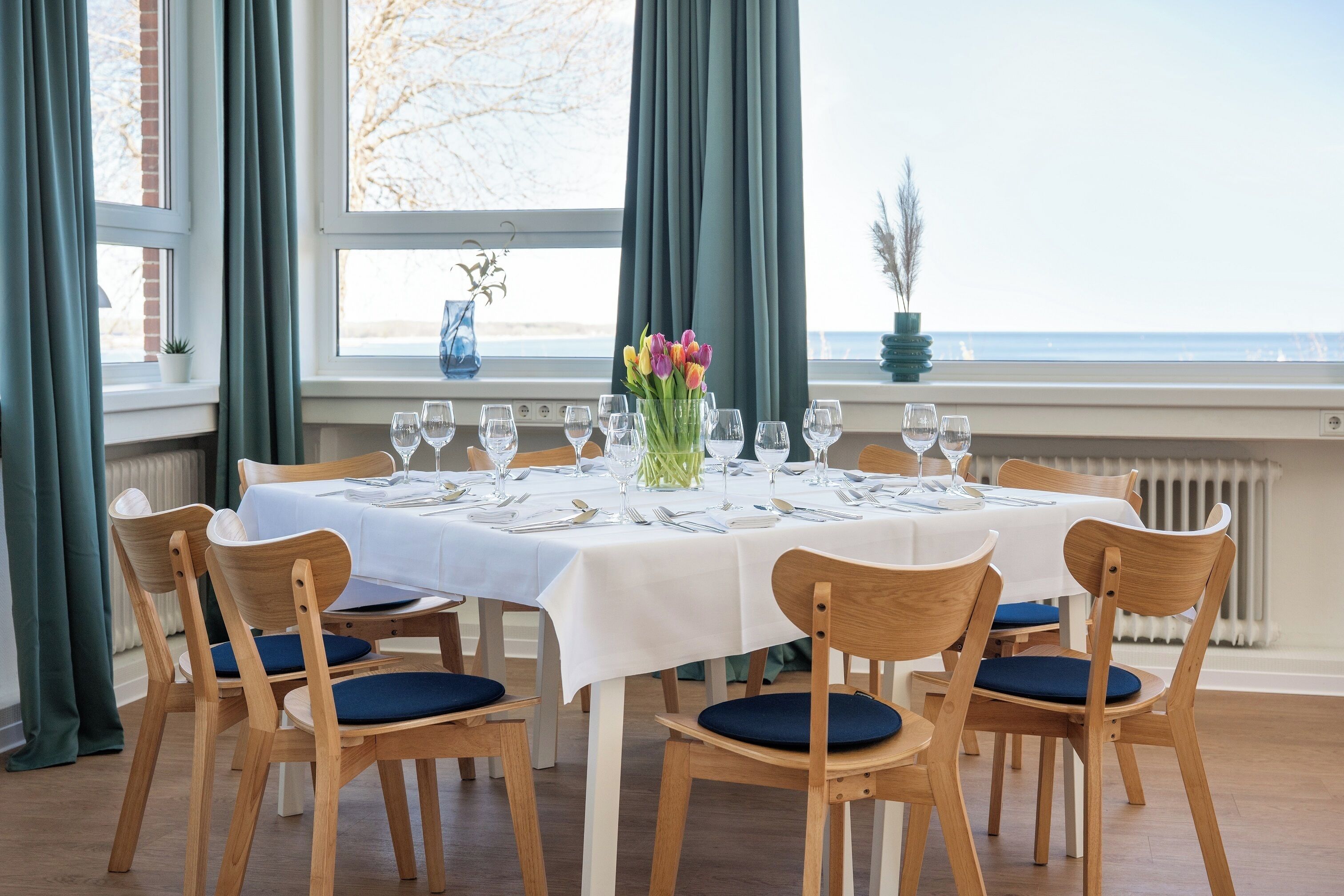 Mesa de comedor preparada para seis personas con sillas, un jarrón de flores y vista al mar.