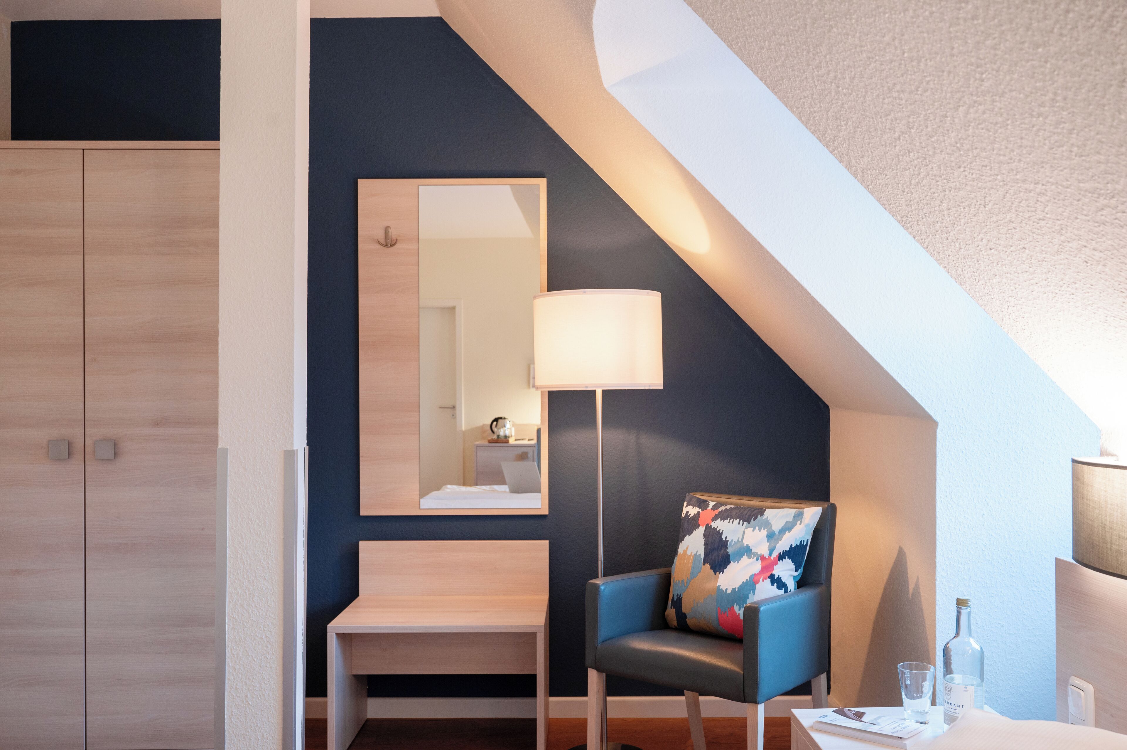 A modern hotel room with a wooden wardrobe, a bench, a chair with a colorful cushion, a desk with a lamp, and a mirror on the wall.
