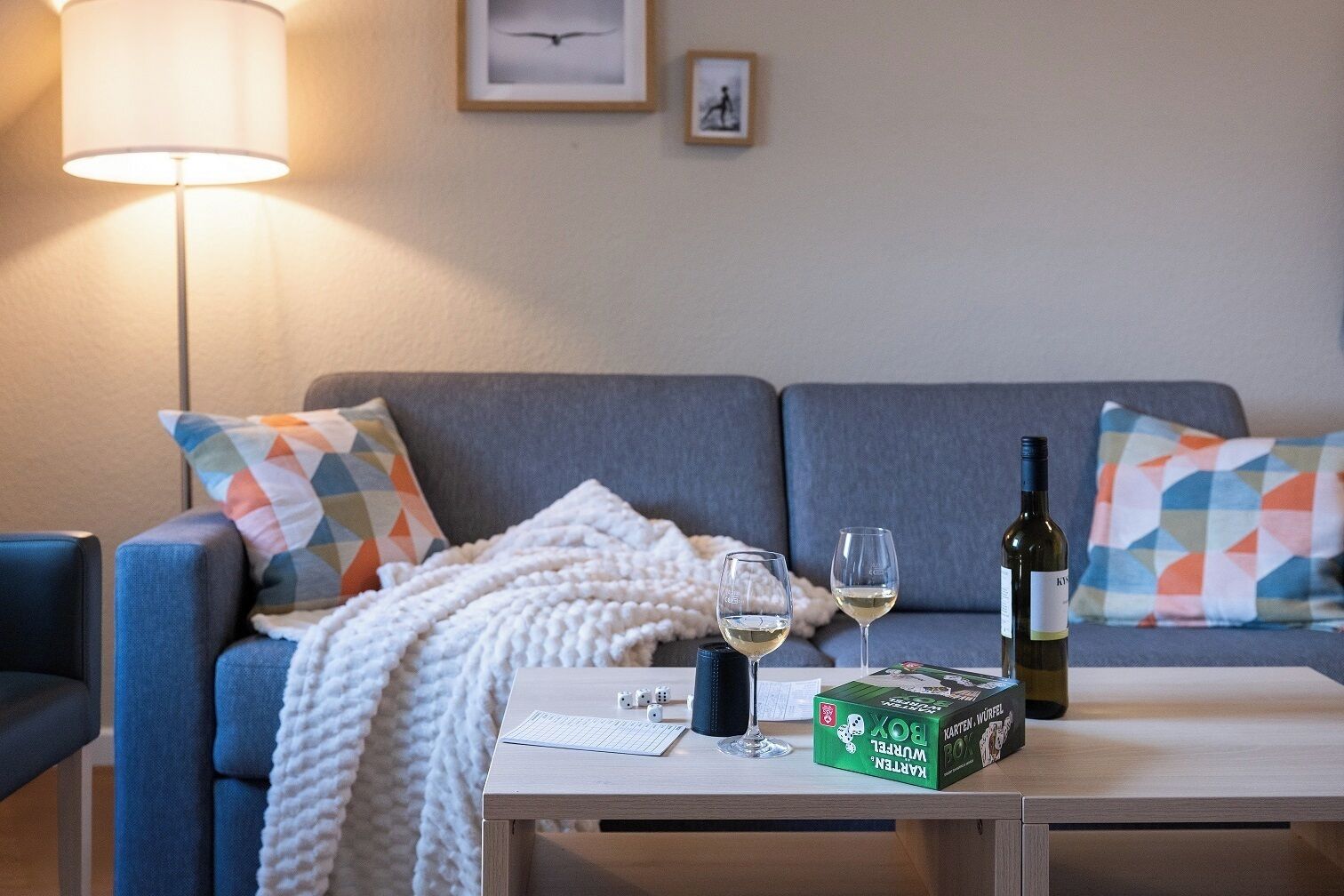 A living room with a sofa, a coffee table, a lamp, two glasses of white wine, a bottle of wine, and a board game box.