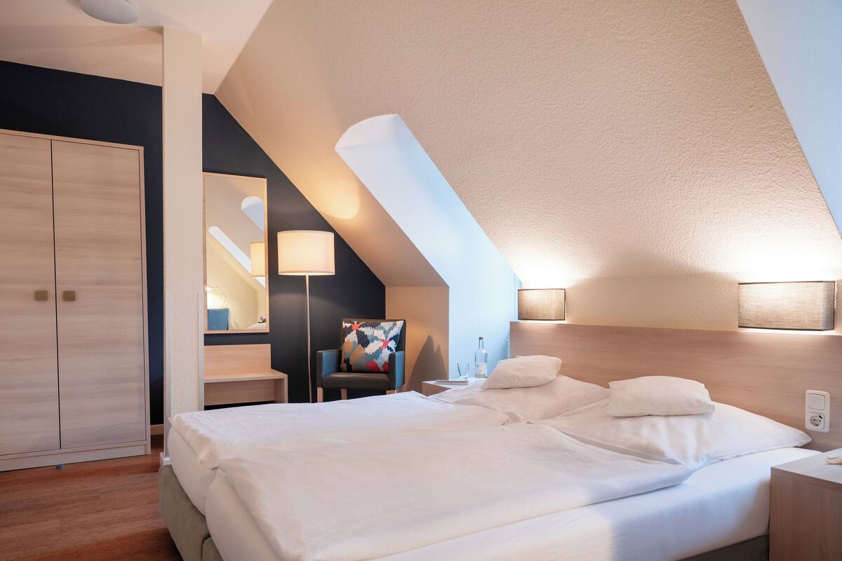 A modern hotel room with a large bed, a desk, a chair, and a wardrobe.