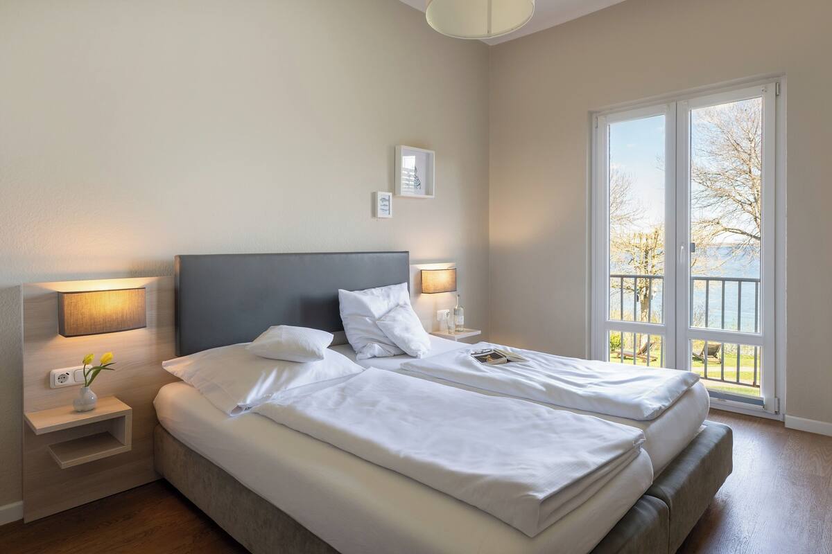 A double bed with white linens, bedside lamps, a nightstand with a vase, and a view of trees through a large window.
