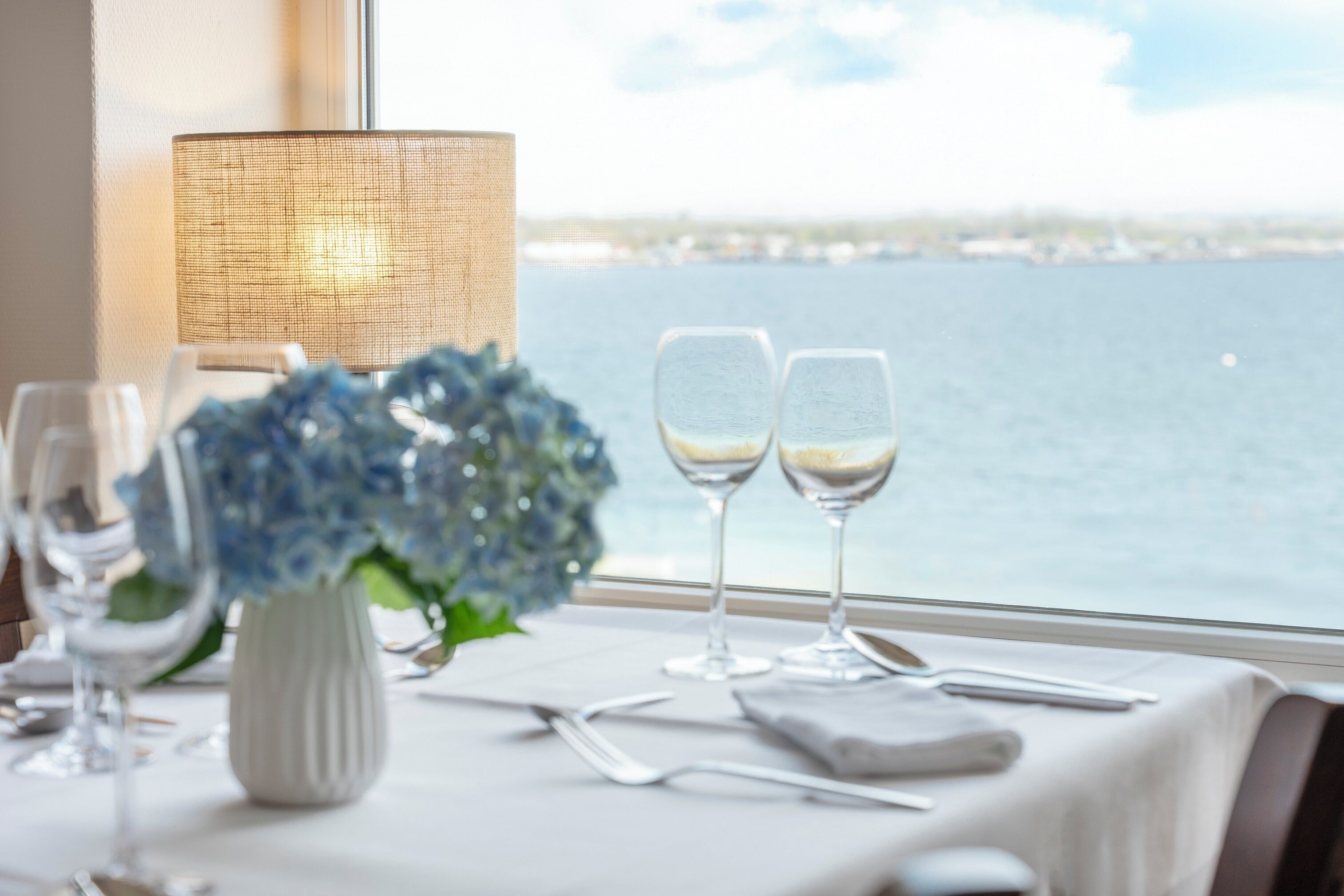 Una mesa dispuesta con copas de vino y un florero, una lámpara y vistas al agua.
