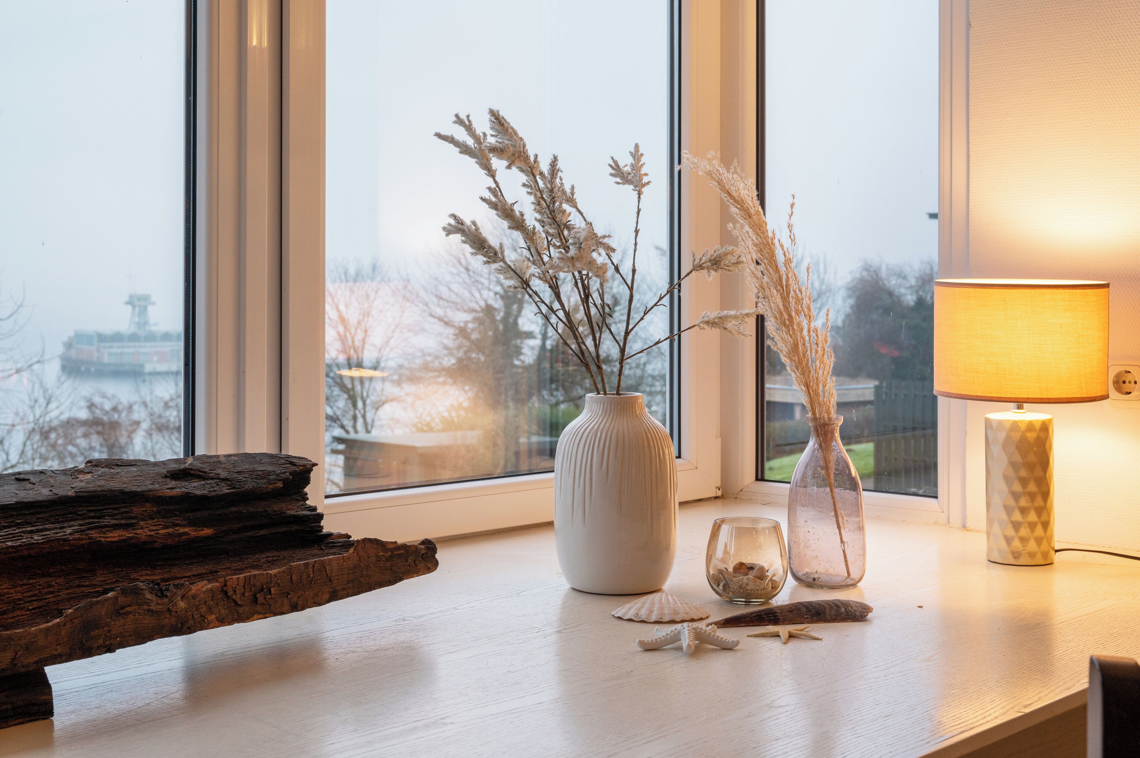 A wooden table with a vase of dried plants, a glass with seashells, and a lamp on a windowsill.