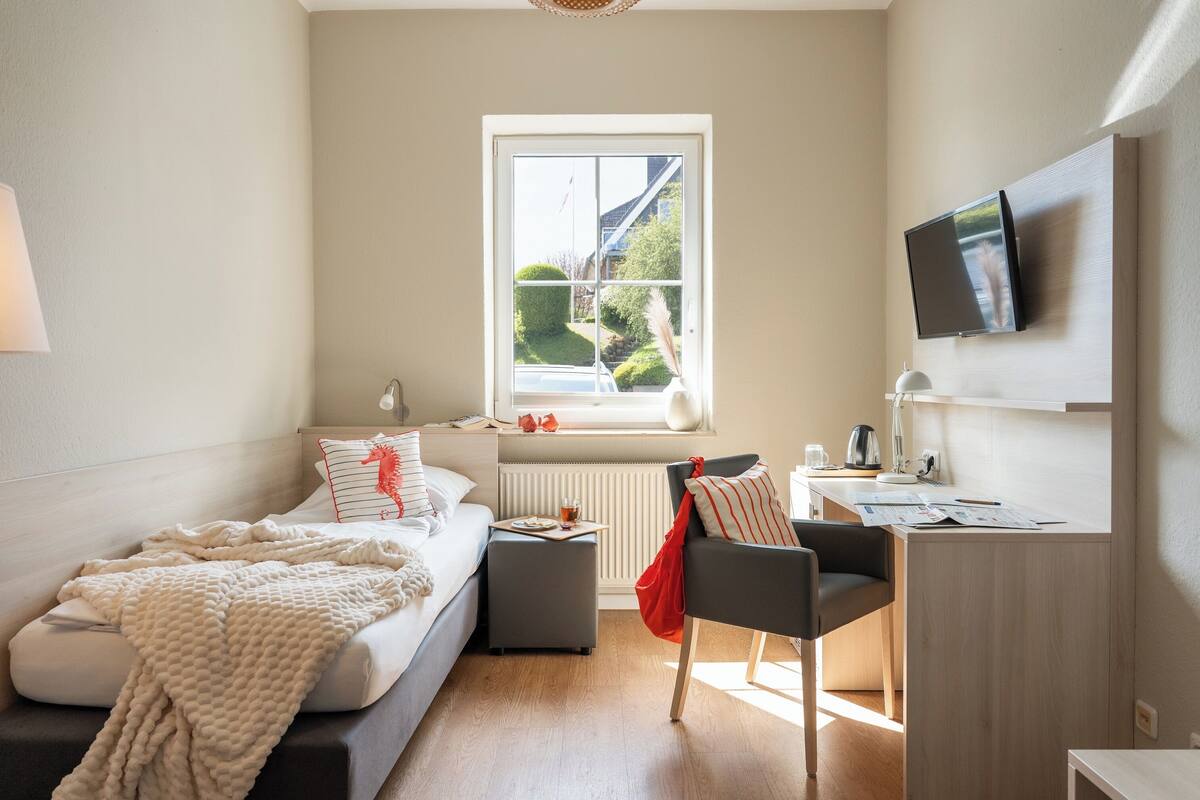 A modern bedroom with a bed, a desk, a chair, a TV, and a window with a view of greenery.