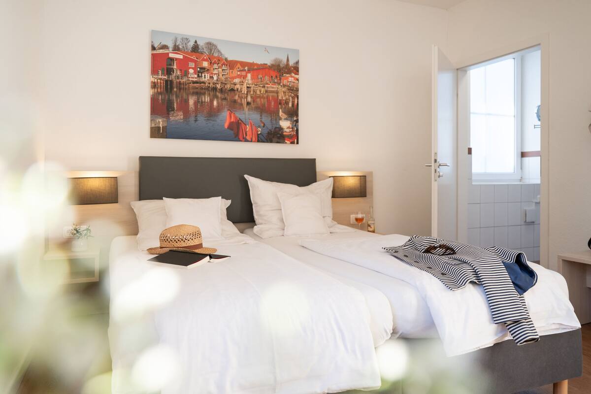 A neatly made bed with white linens, a striped shirt, and a straw hat on the pillows. There is a painting of a red building on the wall above the bed.