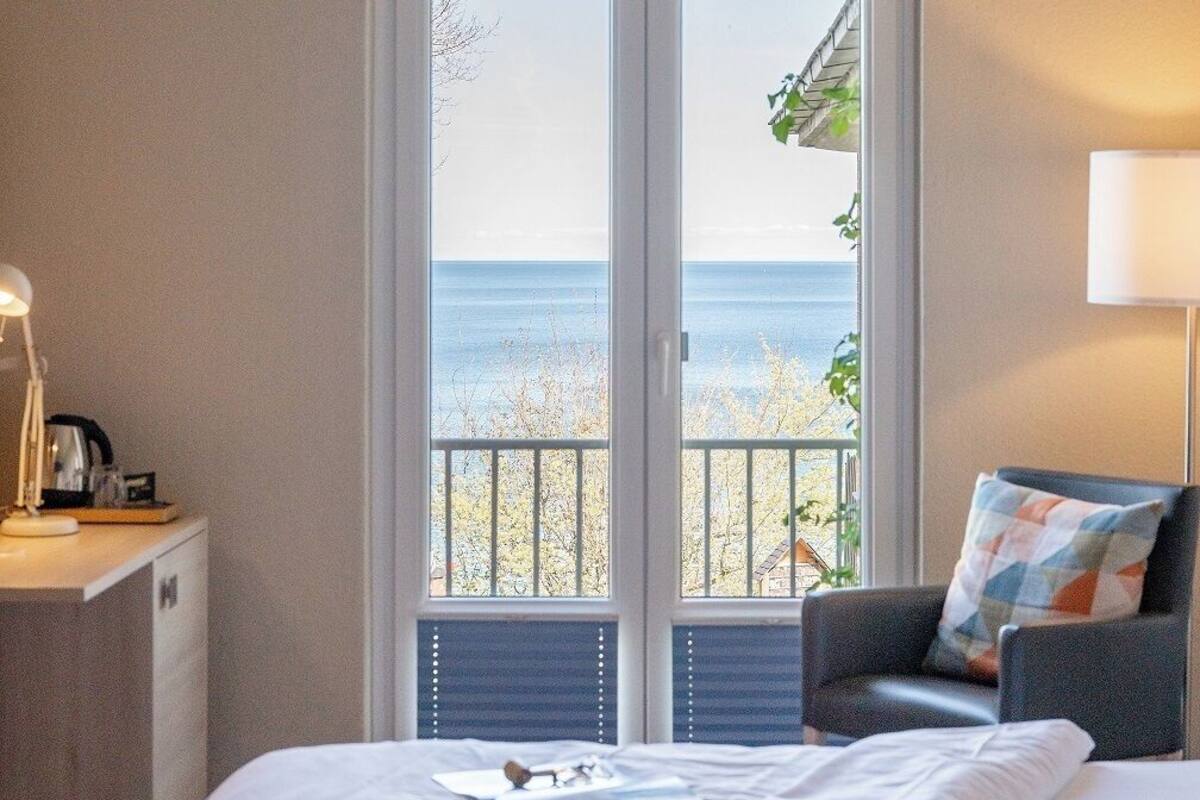 A hotel room with a bed, a desk with a lamp, a chair, and a view of the sea through a glass door.