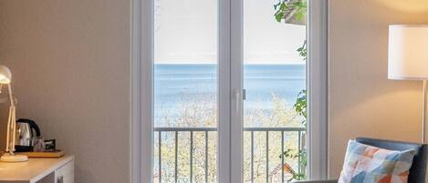 Habitació d'hotel amb un llit, una taula de treball amb una làmpada, una cadira i vista al mar a través d'una porta de vidre.